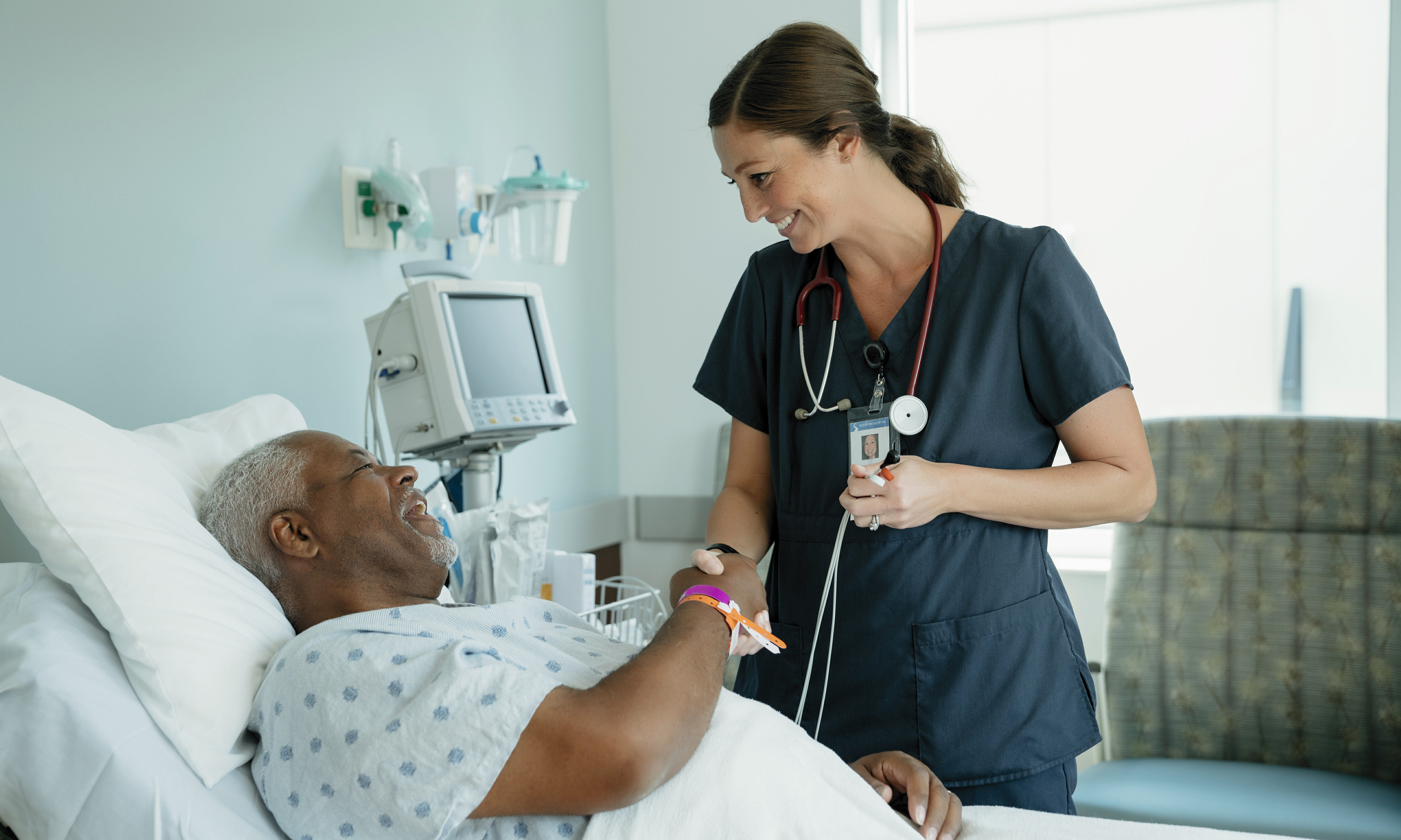 Smiling Nurse Handshaking With Senior Patient Lying On Bed In Hospital